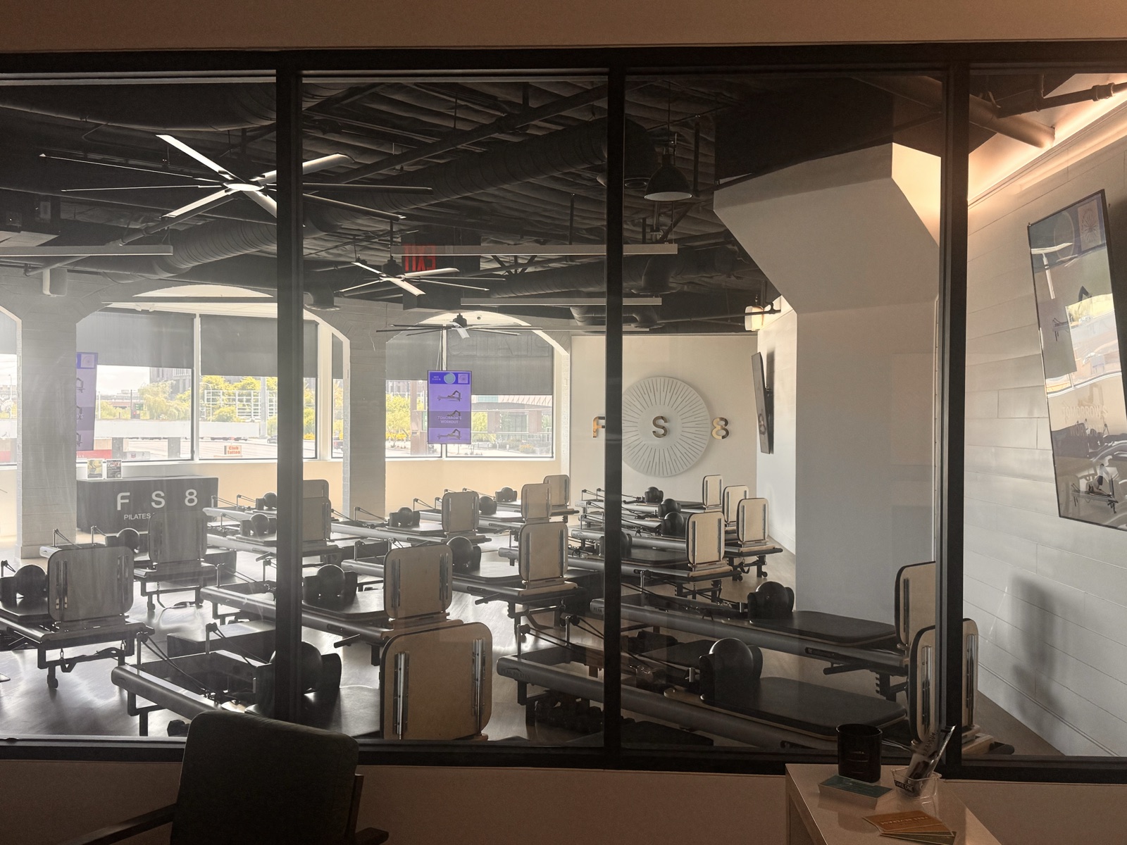After — finished FS8 Tempe reformer studio viewed through the storefront glazing, with rows of reformers, branded logo wall, and warm pendant lighting